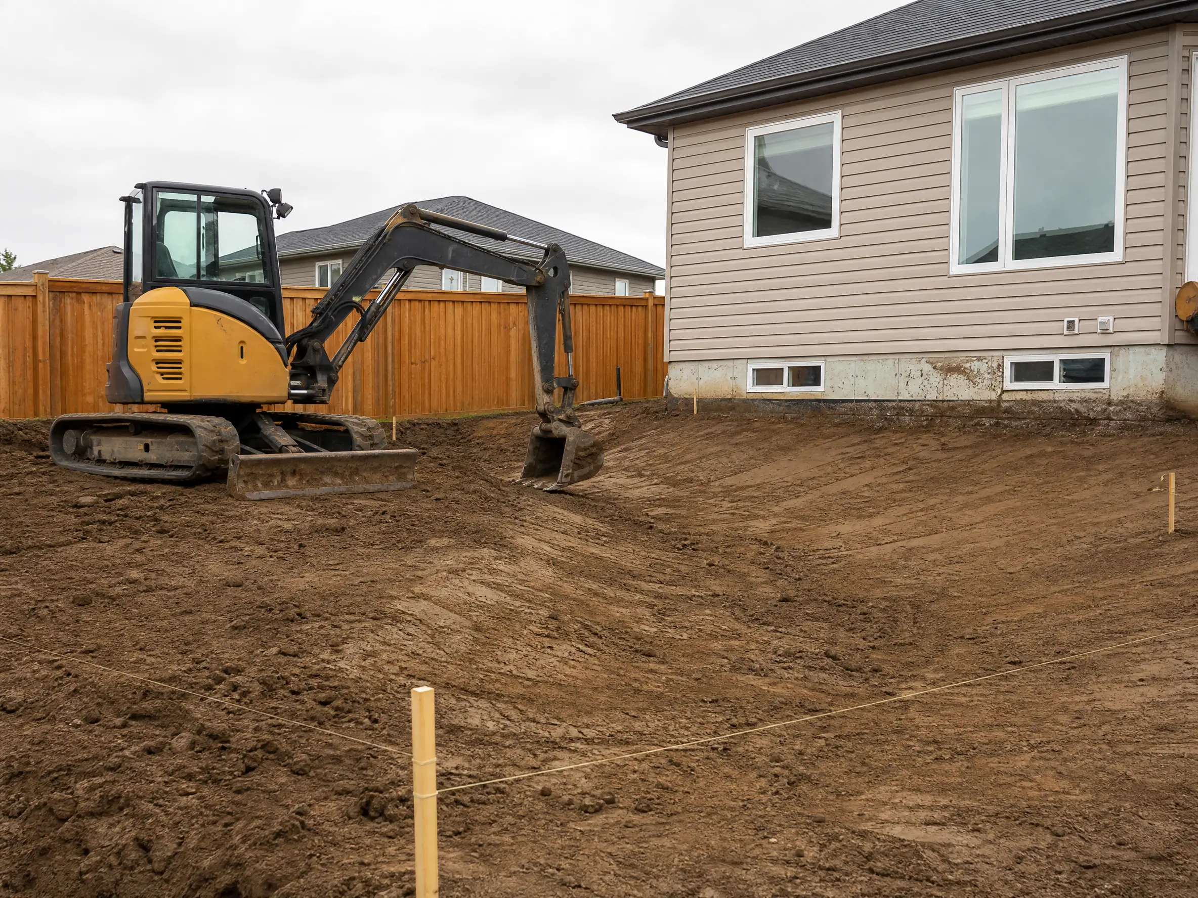A compact excavator regrading a residential back yard toward a swale in Saskatoon, SK